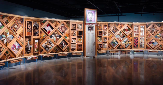 A large and long art installation made of wood frames with pictures and objects woven throughout to look like a large quilt. It is slightly curved and stands in a dark gallery. Photo: CMHR, Jessica Sigurdson.