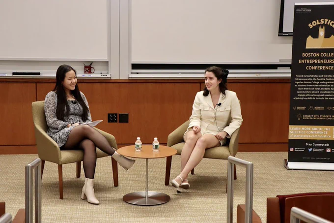 Left to right, me (Maggie Yan) and Meagan Loyst, sitting on muted yellow/green arm chairs  with a small round table in between us with 2 mini water bottles. 

Behind us are two big white boards and a black Solstice conference banner is off to the right side.