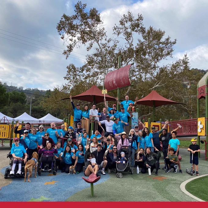 Large group of approximately 40 people in athletic gear on playground jungle gym all making funny faces. Majority of people in photo are wearing blue shirts that read Inclusion Matters. Two people in wheelchairs in the front row and one baby in a stroller and one kid on a scooter.