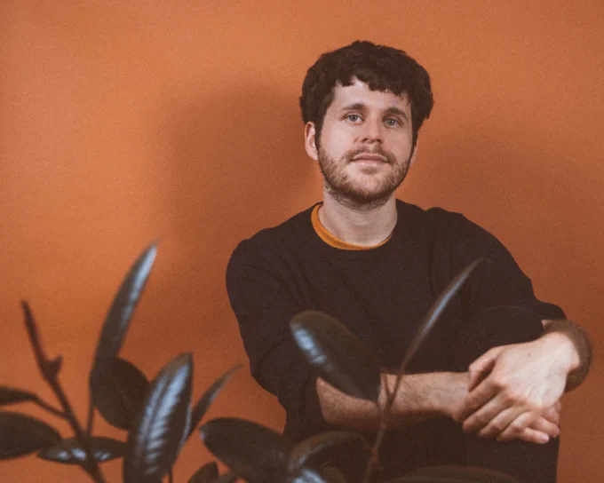 Stephen Becker sits with arms hugging a knee. Smiling and looking at the camera, he's in front of a sienna-painted wall. A plant's leaves are out-of-focus in the foreground.