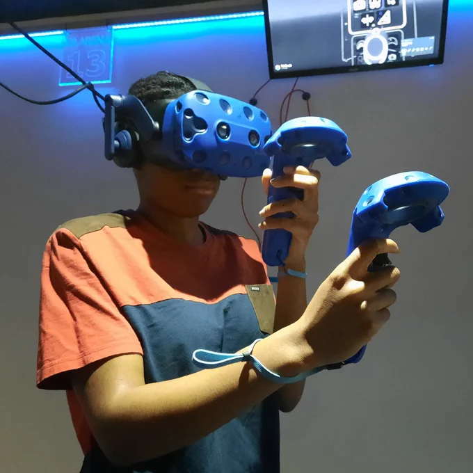 Image of young person using a virtual reality headset. They are holding a controller in both hands. Blue LED lighting is behind them. There is a TV screen showing what they can see inside the headset.