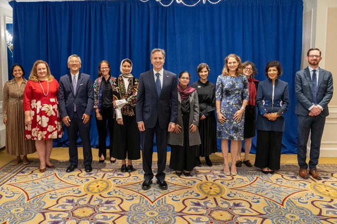 Secretary of State Antony Blinken stands in front of leaders of the State Department’s Alliance for Afghan Women’s Economic Resilience. They are standing on a yellow rug with an elaborate pattern with a blue curtain in the background.