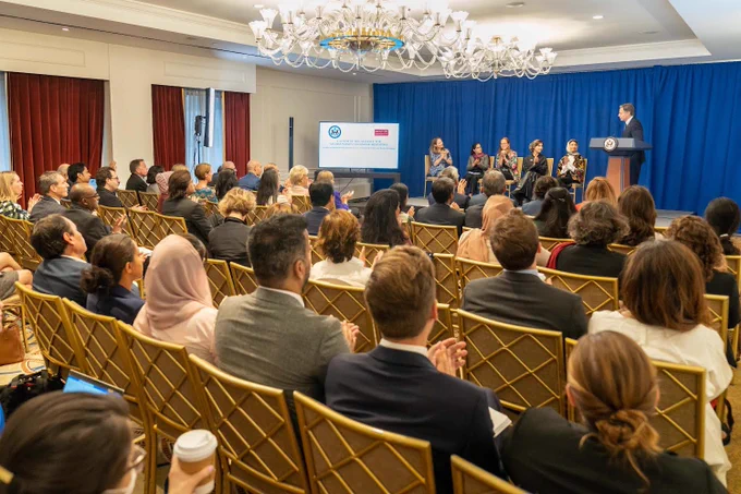 Audience is listening as Secretary of State Antony Blinken stands on a podium with the U.S. Department of State emblem on the middle. Five women are also sitting on the stage as the Secretary announces the State Departments Alliance for Afghan Women’s Economic Resilience.