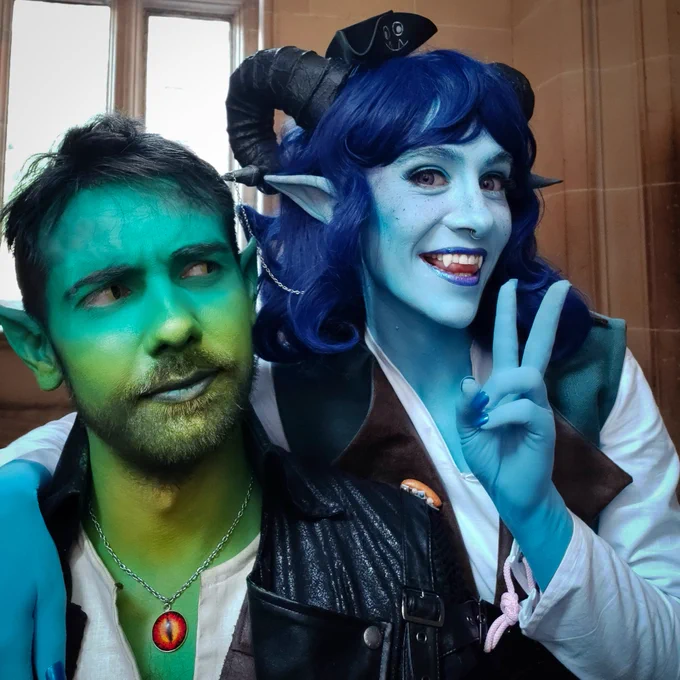 Selfie of me dressed as Jester and Craig as Fjord.

My skin is painted blue, I'm wearing a dark blue wig and have long ears and black ram horns. I'm smiling towards the camera and holding a piece sign.

Craig's skin is painted on a green and yellow ombre. He has pointed ears and is looking at me confused.