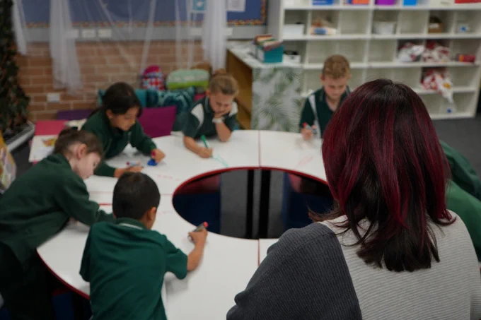 A teacher watching students seated around 4 quarter tables, arranged in a circle. The students are writing on the whiteboard surface of the tables