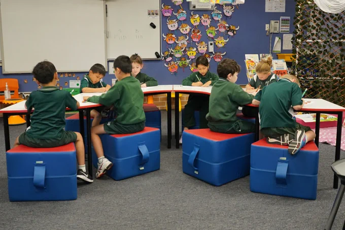 The same students and same 4 quarter tables, this time the tables are arranged in a long curved row. The wedge-shaped red and blue ottomans (with handles) that the students are sitting on are also visible.