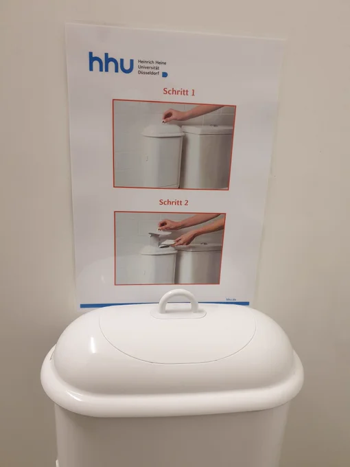 Hygieneeimer im Toilettenraum, darüber bebilderte Anleitung: 1. Mit einer Hand Deckel anheben und festhalten. 2. Mit der anderen Hand Abfall einfüllen.