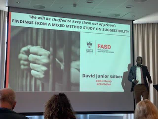 David Junior Gilbert presents the title slide for his presentation entitled, "We will be chuffed to keep them out of prison": findings from a mixed method study on suggestibility. Image on slide includes a pair of hands holdings on to metal bars. 