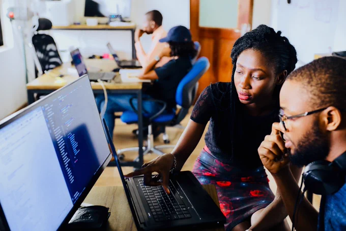 Photo by Desola Lanre-Ologun, on unsplash.com - showing two co-workers talking at a desk and looking at work on a laptop. 