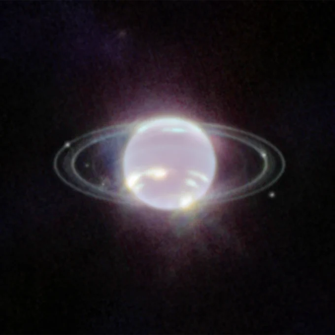 In this Webb image, Neptune resembles a pearl with rings that look like ethereal concentric ovals around it. There are 2 thinner, crisper rings and 2 broader, fainter rings. A few extremely bright patches on the lower half of Neptune represent methane ice clouds. Six tiny white dots, which are six of Neptune’s 14 moons, are scattered among the rings. The background of the image is black.
