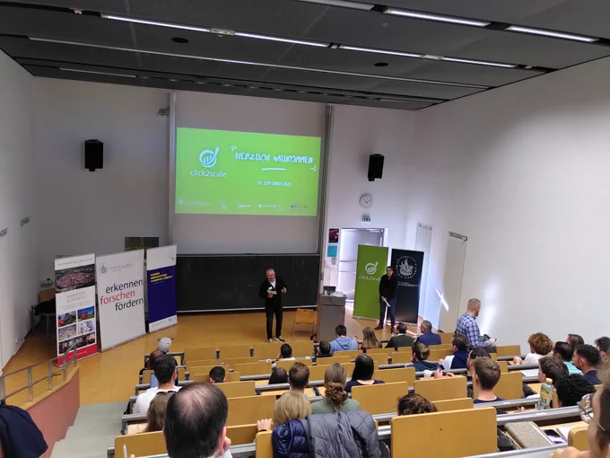 Menschen in einem Hörsaal vor einer Leinwand. Ein Referent steht vor der Leinwand, der zum Publikum spricht.