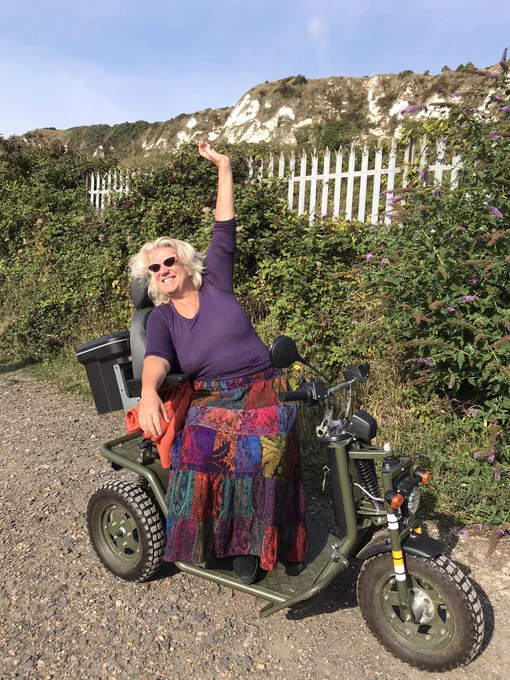 Katy is raising an arm in triumph and beaming broadly. Her motor scooter is in army green; she is wearing a purple too and brightly multicoloured patchwork skirt. She is by brambles and buddleia, with chalk cliffs and a blue sky behind her.