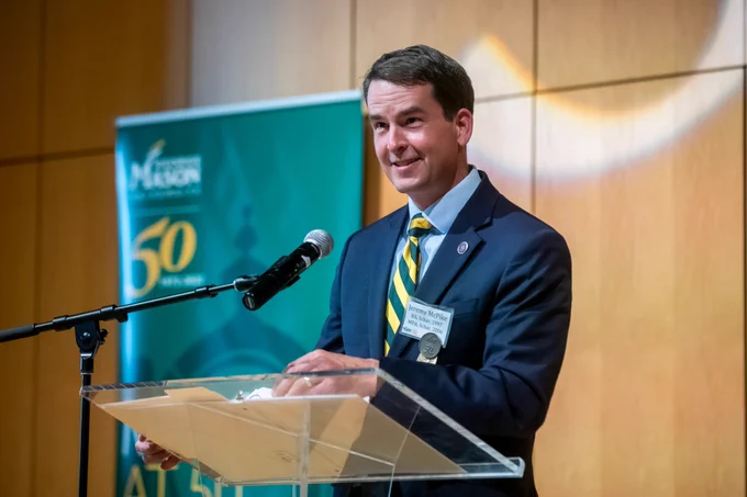 Mason alumnus and State Senator Jeremy McPike speaking at a podium. He is wearing a navy blue suit with a green and gold striped tie.