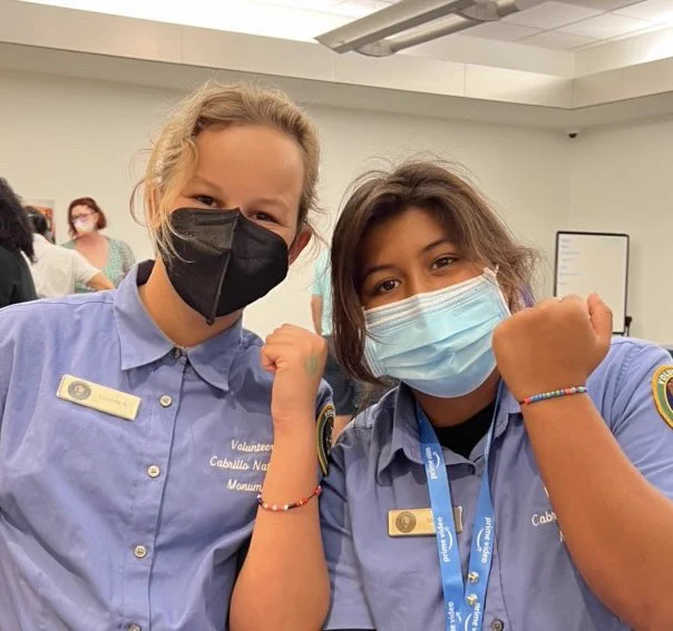 Two girls wearing face masks and blue “volunteer” shirts with name badges show off their binary code bracelets at a STEAM (science, technology, engineering, art, and math) fair. 