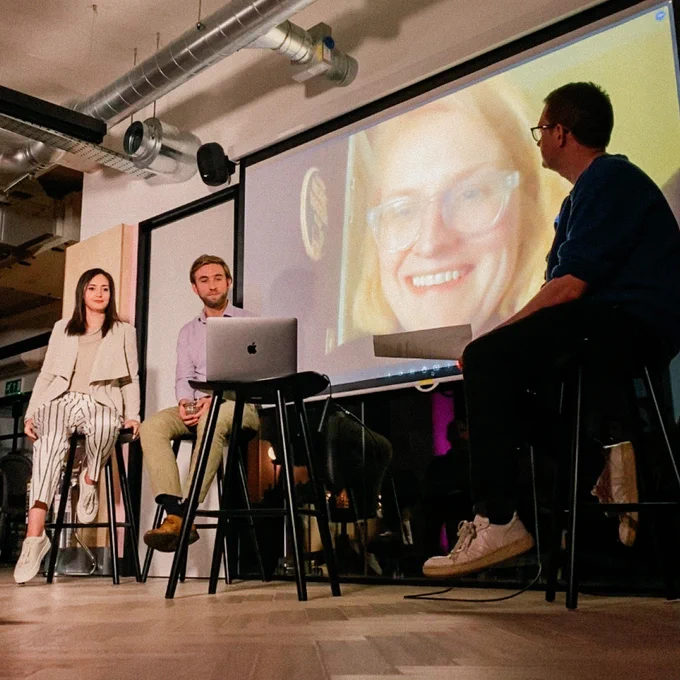 A shot of our event in action in our studio. Two of our panellists, Irina and Lewis, are sitting on stools beside a projector screen — our final panellist, Sarah, is visible on the screen, talking and smiling. Our moderator is sitting opposite them.