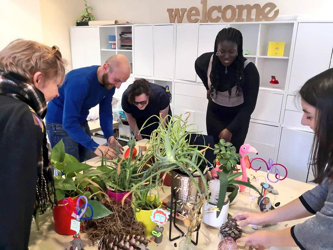fovea people gathered round and leaning over a table of leaves plants and clay. Wooden words spelling 'welcome' in background 