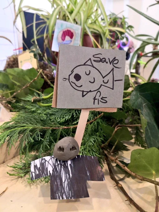greenery witn a tony placard held by a clay person. Placard has drawing of a fish witn words 'save fis'