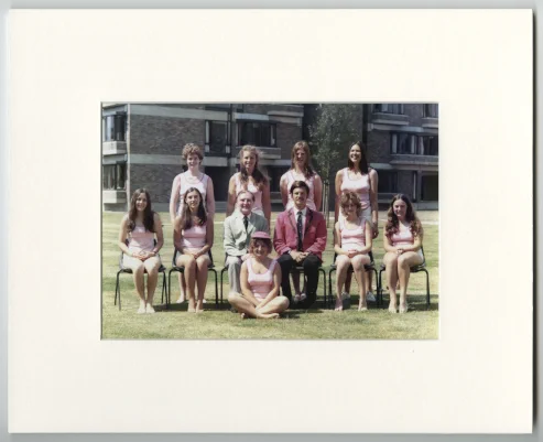 Nine women dressed in pink rowing tops are posing for a sports team photo with their two male rowing coaches. Four women are standing behind a row of chairs and two men, one of them is Canon Duckworth, are sitting in the centre of the picture with two women seated on either side of them. There is another woman wearing a pink cap sitting cross-legged on the grass at the front of the picture.