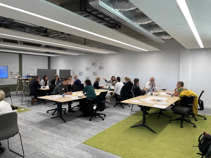 A photograph of the whole room with groups sat around tables discussing ideas