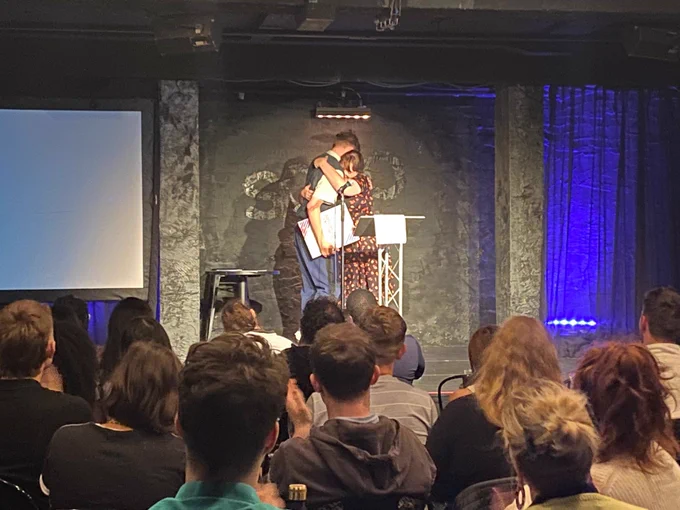 Photograph of Sam Grabiner on stage accepting his award. He is stood in front of an audience, hugging the announcer.