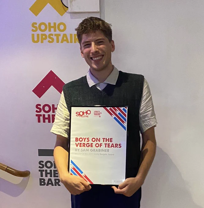 Photograph of Sam Grabiner, a young white man stood in the foyer of Soho Theatre. He is smiling, wearing a white short sleeved shirt, blue jeans and a grey sweater vest. He is holding a large award which reads Boys on the verge of tears by Sam Grabiner