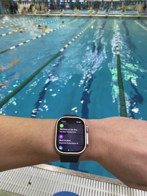 In the foreground a wrist with an Apple Watch Ultra on it with the MySwimPro app on the screen. In the background is an indoor swimming pool with lane lines.