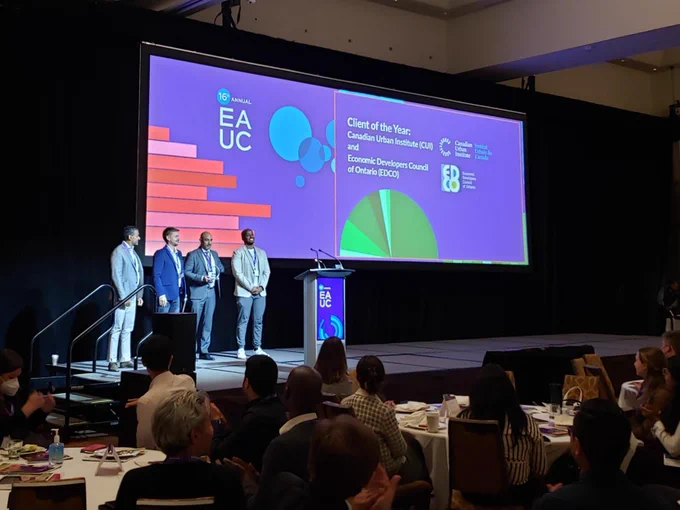 Four people are on stage for an award reception. The stage screen reads "Client of the Year: Canadian Urban Institute (CUI) and Economics Developers Council of Ontario". 