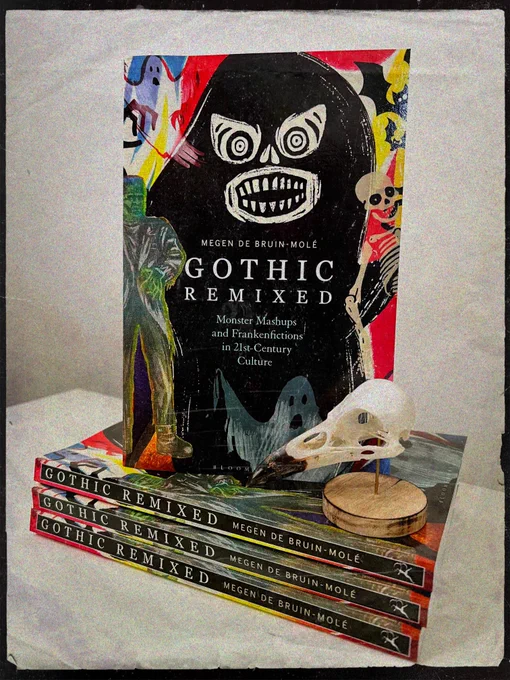 Four copies of the book Gothic Remixed: Monster Mashups and Frankenfictions in 21st-Century Culture stacked on top of each other, with the top copy stood upright so you can see the ghosts, skeletons, and Frankenstein’s monster on its cover. On top of the book pile next to the cover is a crow’s skull on a wooden stand.