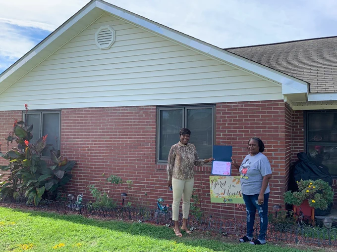 Yard of the Month Winner Sandra McKnight is standing outside her brick front home and is receiving her certificate from HACG Staff member Sharita Oates.
