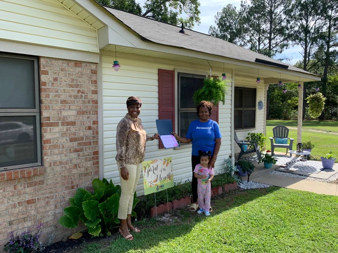 Yard of the Month Winner Roasa Rawls is standing with a little girl outside her home and is receiving her certificate from HACG Staff member Sharita Oates.