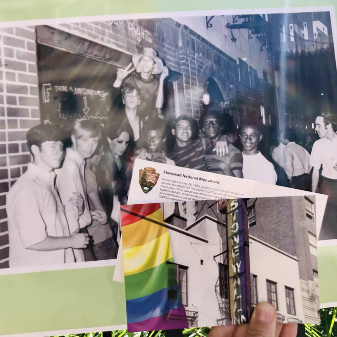 NPS/Burna image: Two postcards featuring a stylized historic photo of the Stonewall Inn and National Park Service logo are held up in front of stop number 4 on the Stonewall National Monument Fence Exhibit. This image highlights an unidentified group of young people posing for photographer Fred W. McDarrah outside the boarded-up Stonewall Inn after the uprising.