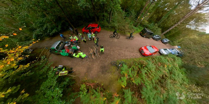 A small group of people standing on a dirt road in a forest, around a pickup truck, receiving a safety briefing. Several mountain bikers ride past along the dirt road, and some cars are parked nearby.