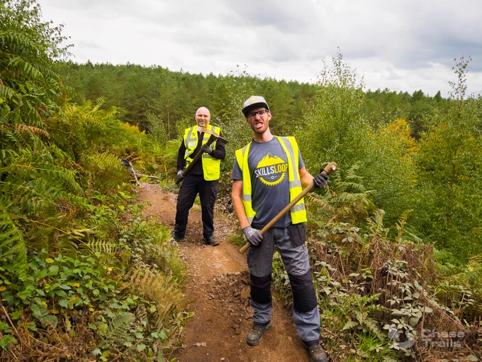 Two volunteers standing on the trail holding hand tools and pulling a face at the camera.