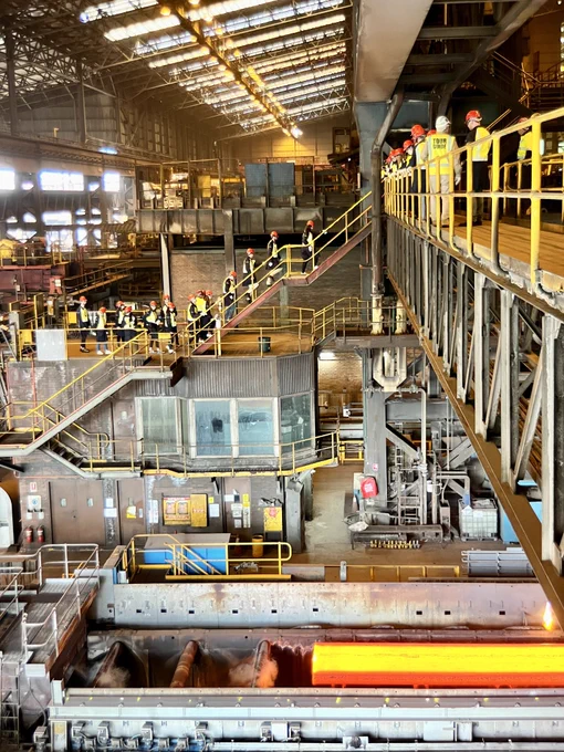 A tour group ascends stairs and a walkway over the slab caster, with a glowing slab of hot steel on a conveyor belt below.