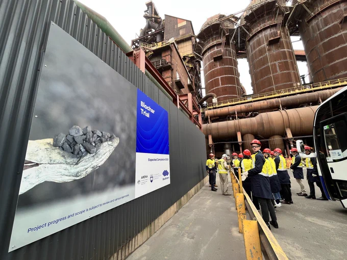 A tour group stands in front of a billboard that displays a 'Biochar trial' title and two hands in gloves, holding material, with the stoves of the blast furnace behind the group in the background.