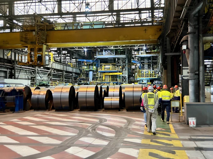 A tour group walks past steel coils in a warehouse.