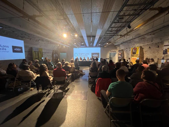 Image shows the audience, looking forward to the panel of guests speakers. There is a large white screen behind the speakers which says: Sight Loss Councils, with a small black box to the side which says "RIP Her Majesty."