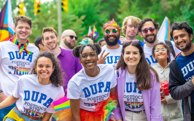 A group of people wearing DUPA postdocs with pride t-shirts smile at the pride parade in Durham