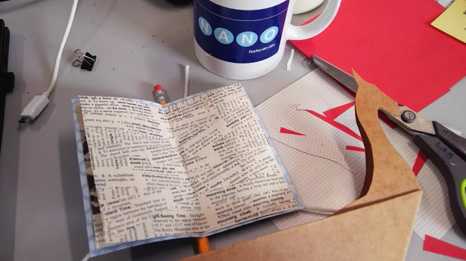 close up of a messy desk wiht a 'NANO' mug, scissors, and part of a paper viking longship, wiht a sail made out of old book pages