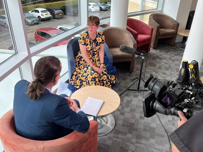 Penny is sitting facing a window while she is interviewed. We can see the back of the interviewers head and the camera. 