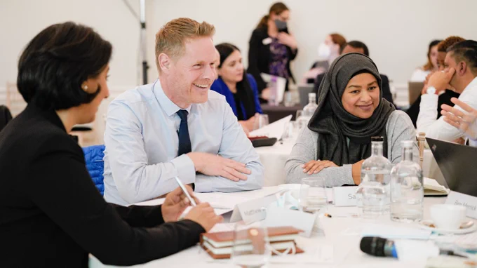 People sit around a table in an Leadership College event smiling and laughing. A man wearing a light blue shirt and a tie and a woman wearing a grey hijab are in focus. In the background several other people are sitting round their tables in discussion.