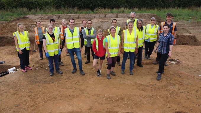 Group of 13 volunteers and 4 staff wearing hi vis and standing in Trench 3.