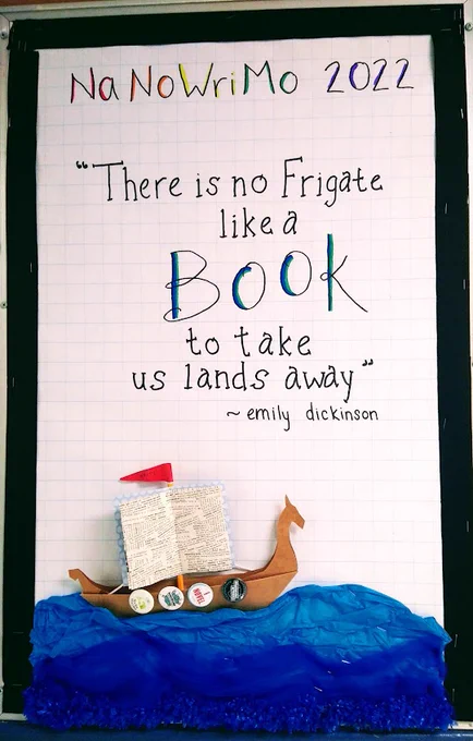bulletin board edged in black, with the quote "there is no frigate like a book to take us lands away" by emilyi dickinson. There is an image of a viking longship with a sail made from upcycled book pages, and #nanowrimo buttons as shield, sailing on a blue tissue paper sea.
