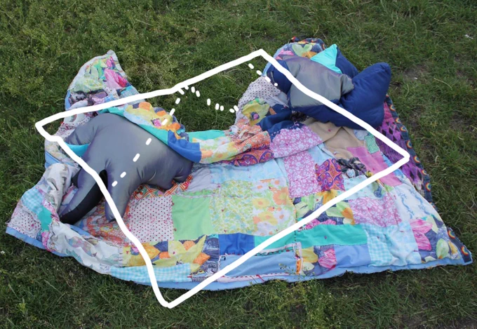 On a grassy ground, a blanket patched together in big stitches and from colourful fabrics holds two pillow objects that rest gently on and under the blanket’s patterns. Above, and shifted slightly diagonally is a simplified white outline illustration of the same blanket. 