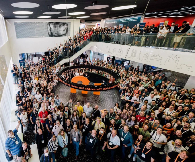 A big room full of MoodleMoot Global attendees, in a shot taken from above. Everyone is looking at the camera and smiling