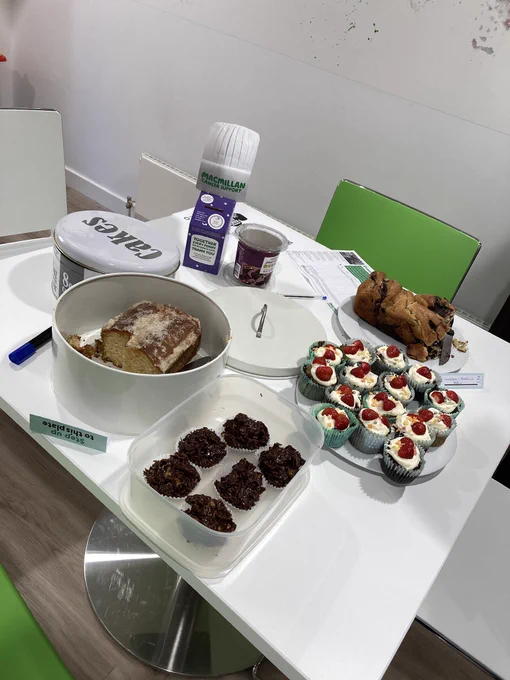 A white square table with a chocolate babka loaf, a lemon drizzle loaf cake, chocolate cornflake cakes and chocolate cupcakes with strawberries and a donation box for macmillan cancer care