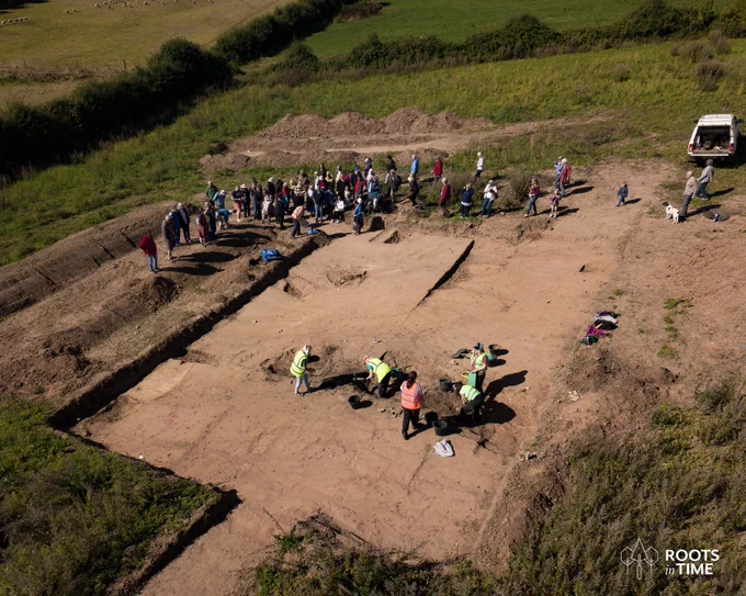 Aerial view of excavation area with four volunteers digging a ditch and a large tour group arriving.