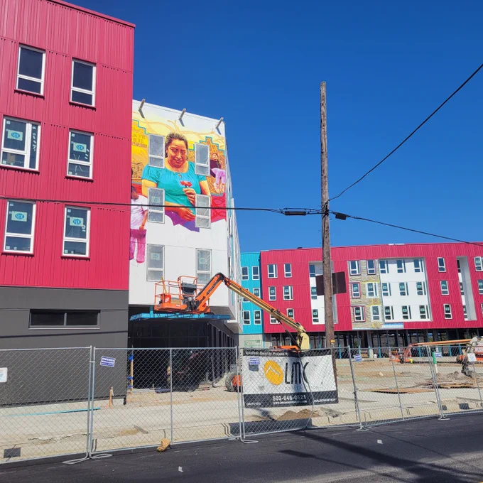 Image of Las Adelitas - four story building, pink and white, with windows - still under construction. This picture includes a partially completed mural of a woman