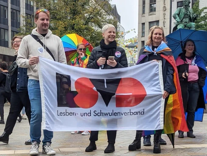 Auf dem Bild sind drei Menschen mit einer Flagge zu sehen. Auf der Flagge sind die vier Buchstaben LSVD zu lesen. LSVD steht für Lesben- und Schwulenverband. Die linke Person trägt eine Blumenkrone. 