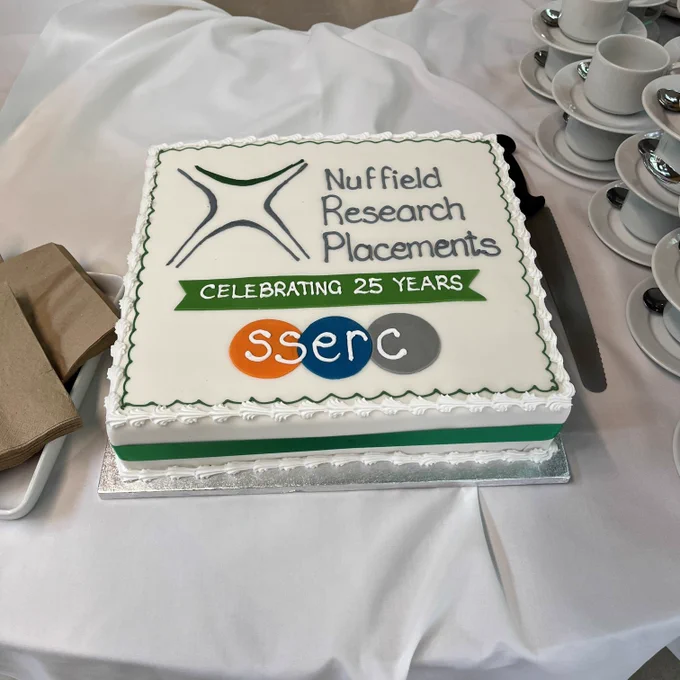 Large square cake with white icing that has the green and grey triangular NRP logo and circular SSERC logo on it and says Nuffield Research Placements, celebrating 25 years